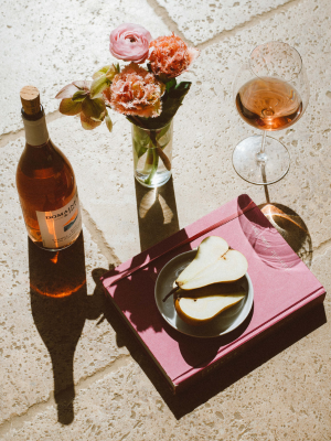 A bottle of rosé wine beside a glass of wine, with a small plate of sliced pear on a pink surface, accompanied by a vase of fresh roses.
