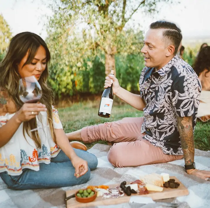 two people, a man in a black and hole floral shirt and a woman holding a wine glass, are sitting on a blanket on the grass as the man holds a bottle of Domaine Roy wines up