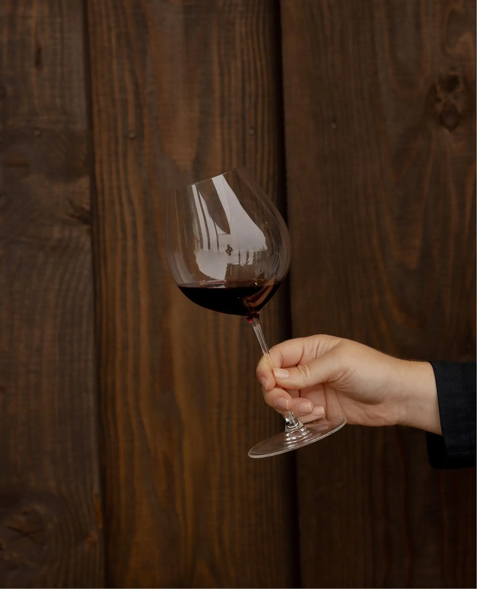 Hand holding a wine glass filled with red wine against a rustic wooden background, emphasizing the elegance of wine tasting and enjoyment.
