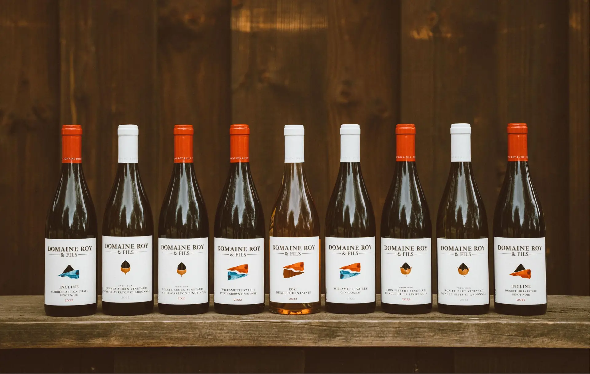 A selection of eight wine bottles displayed on a wooden shelf, featuring various labels and colors, showcasing a range of white wines with distinctive caps and branding.