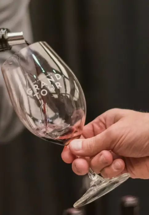 A hand pouring red wine into a branded Grand Tour wine glass, showcasing the elegant design and rich color of the wine against a blurred background.