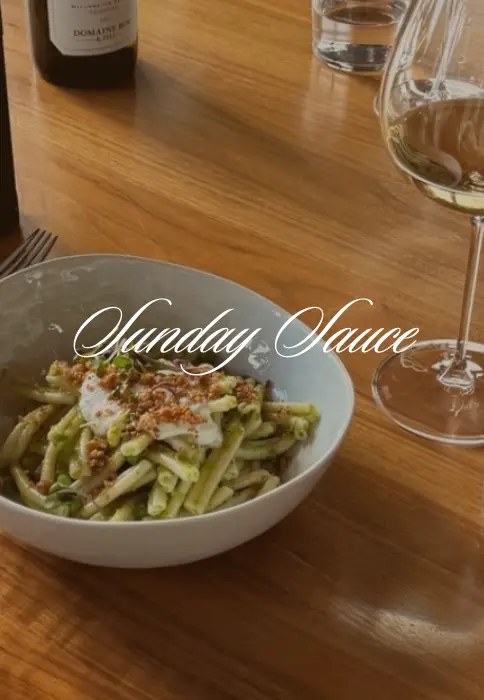 A bowl of pasta with Sunday sauce topped with creamy ricotta and crispy breadcrumbs, accompanied by a glass of white wine, set on a wooden table.